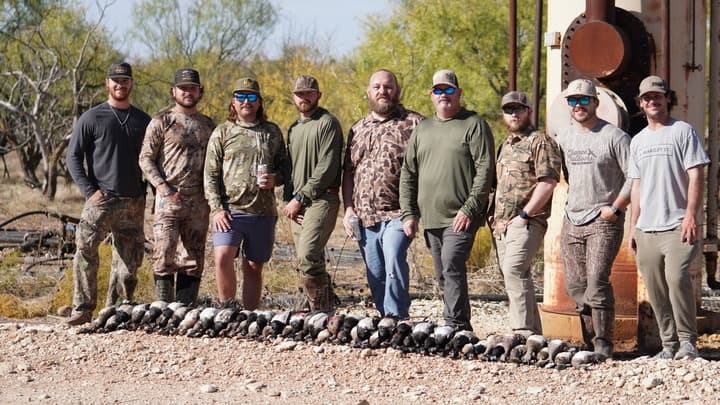 Nine hunters posing behind a large harvest of ducks laid out on the ground outdoors.