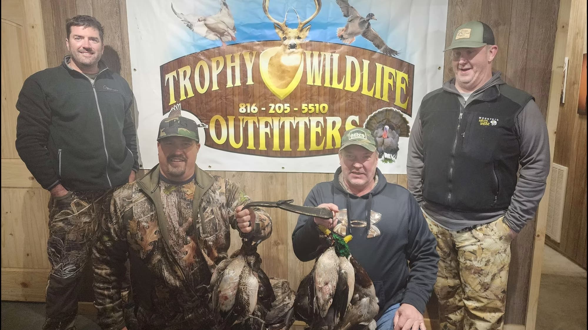 Four men pose with harvested ducks in front of a Trophy Wildlife Outfitters banner.