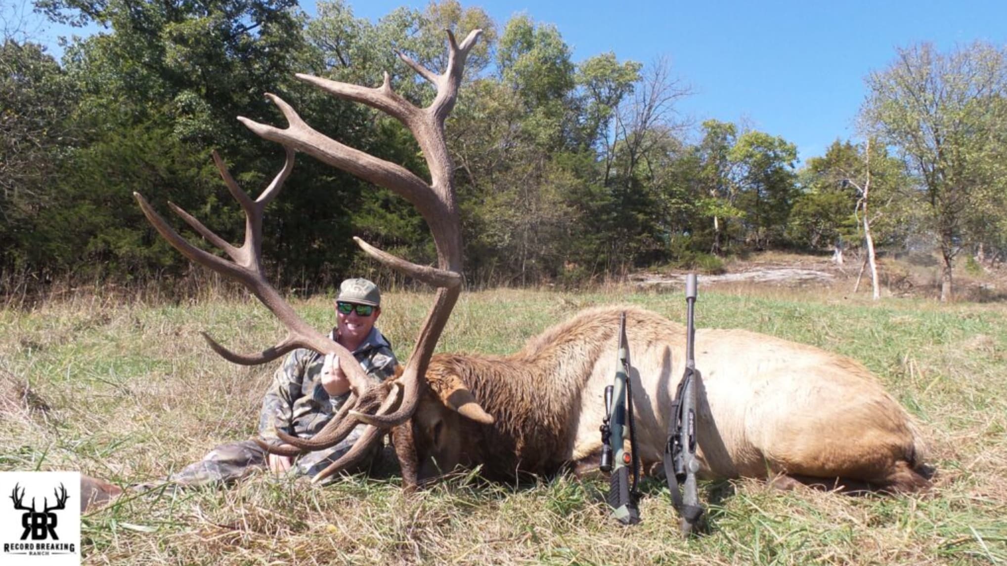 A smiling hunter poses behind a harvested bull elk with massive antlers, two rifles leaned against it.