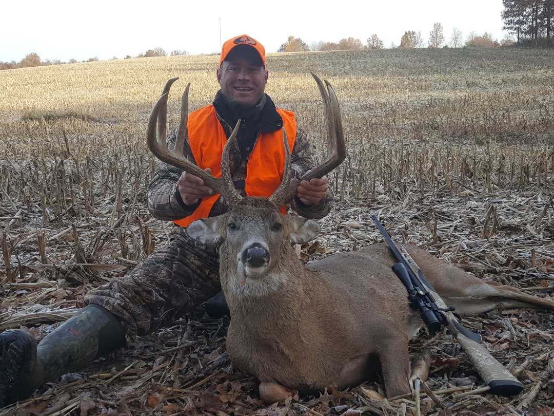 Hunter in orange vest holds large buck antlers beside downed deer with rifle in a harvested field.