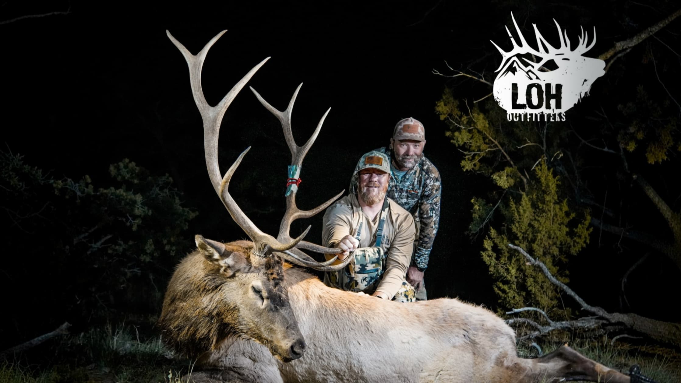 Two hunters pose proudly behind a large bull elk with majestic antlers at night, with the LOH Outfitters logo in the top r...