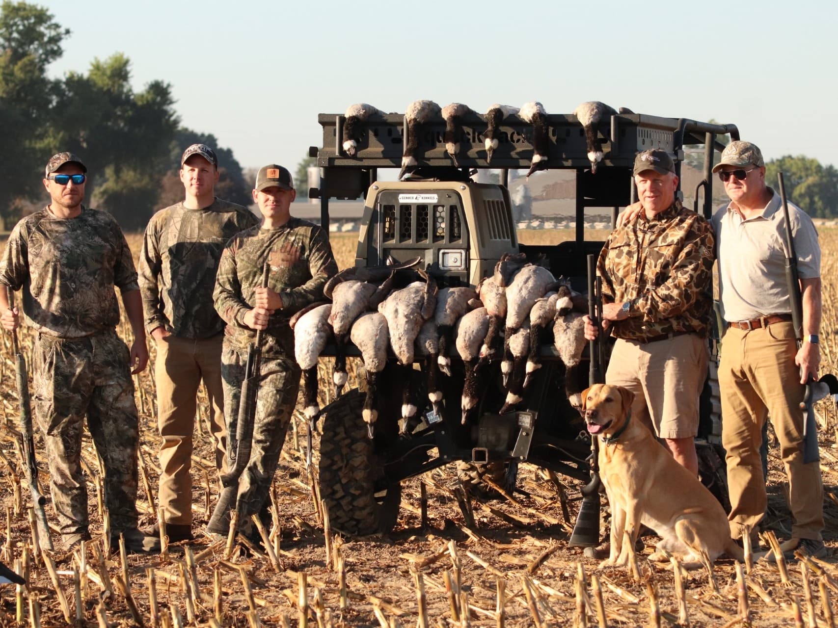 Group of hunters pose with a vehicle carrying many geese. A dog sits near them. Hunters wear camouflage and hold firearms.