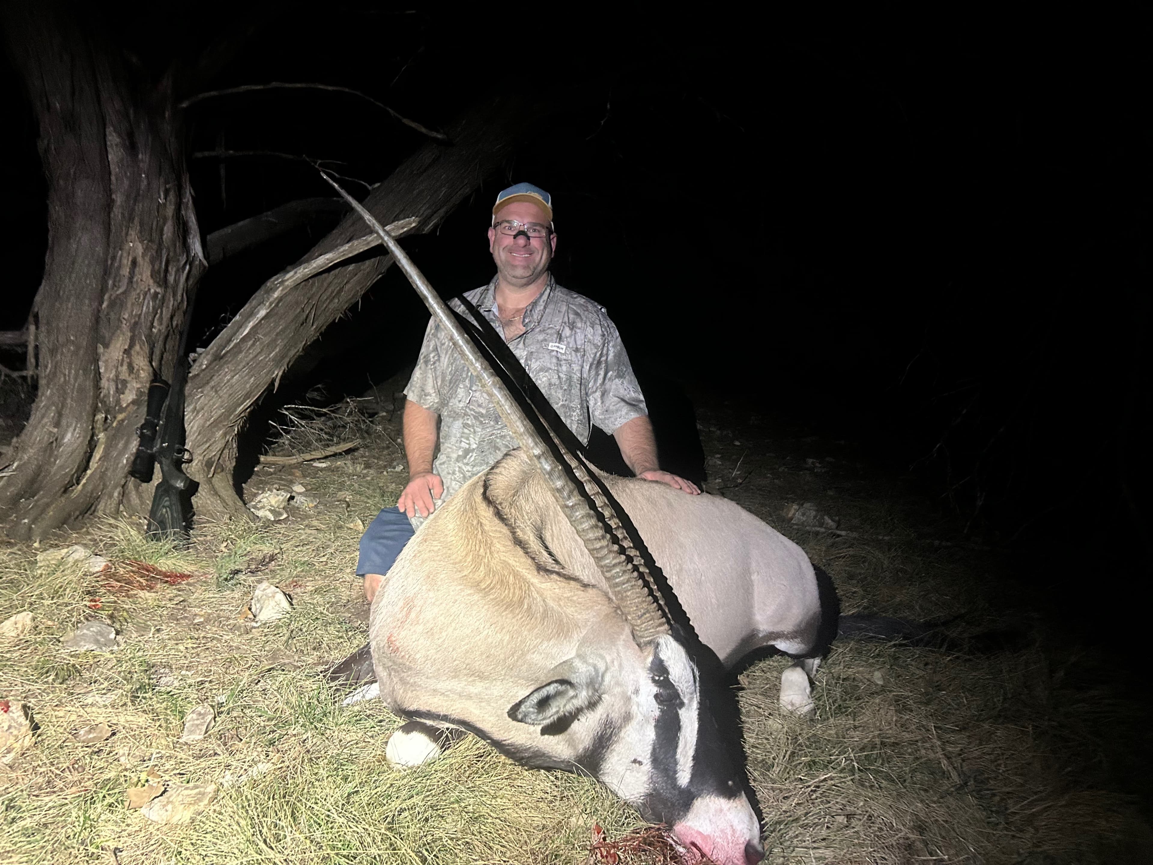 Man poses with dead oryx. The oryx lies on the ground with large horns. A rifle leans against a tree. Nighttime outdoors.