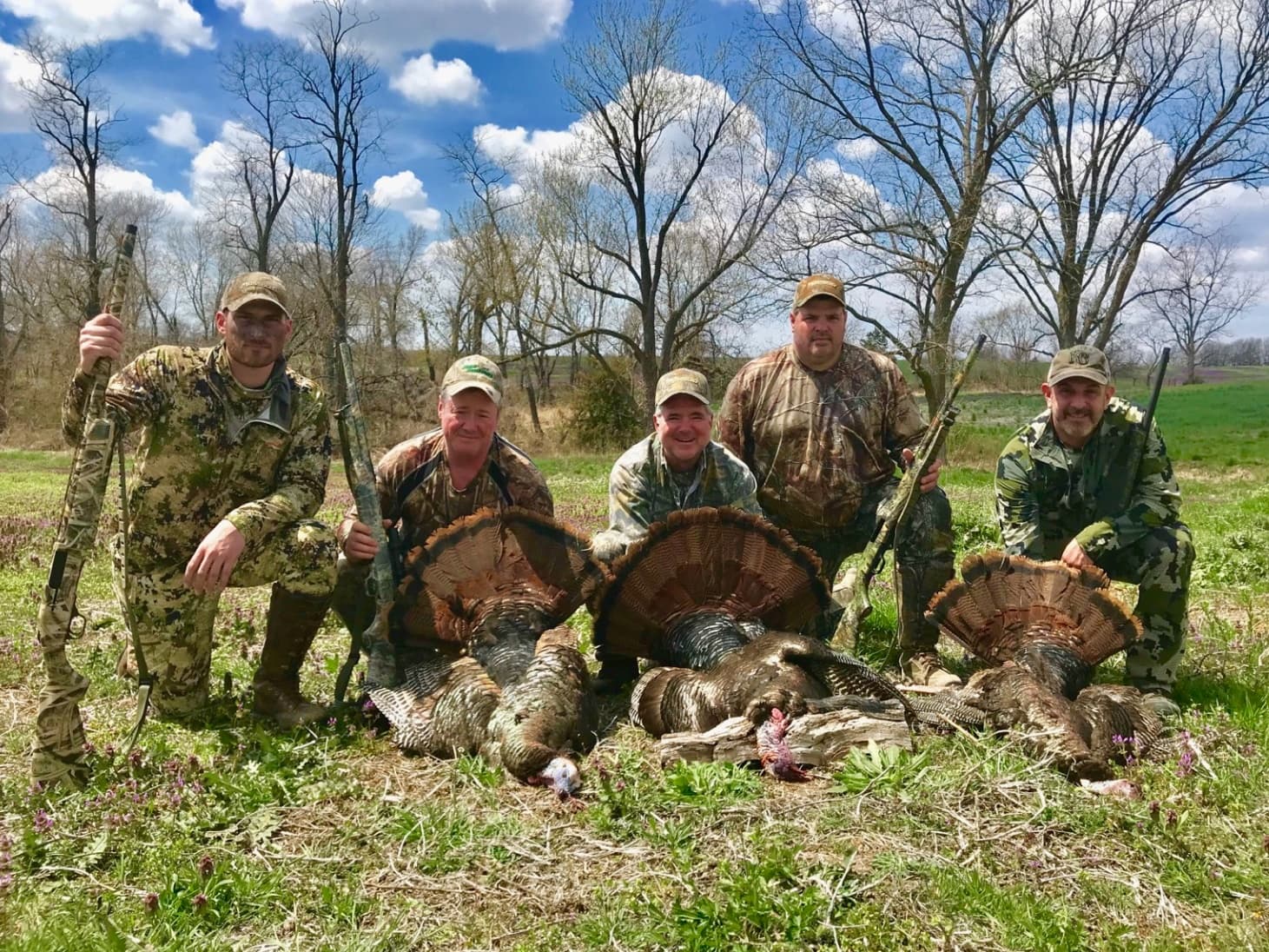Five hunters kneel with shotguns, posing with four harvested turkeys. Hunters wear camouflage in a grassy field, sky with ...