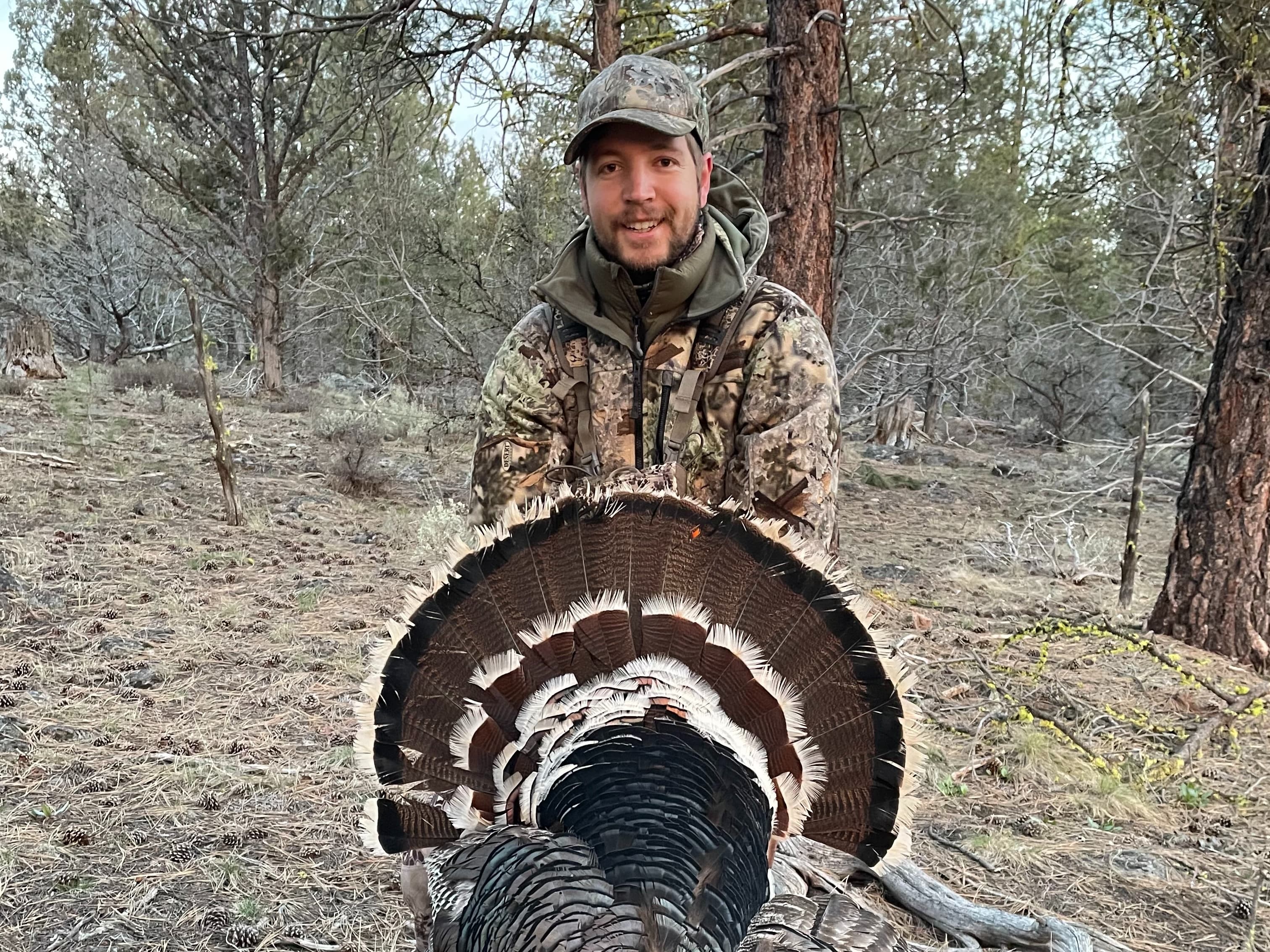 Man in camouflage gear smiles, holding a large, fanned-out turkey tail. Forest backdrop with trees and dry brush.